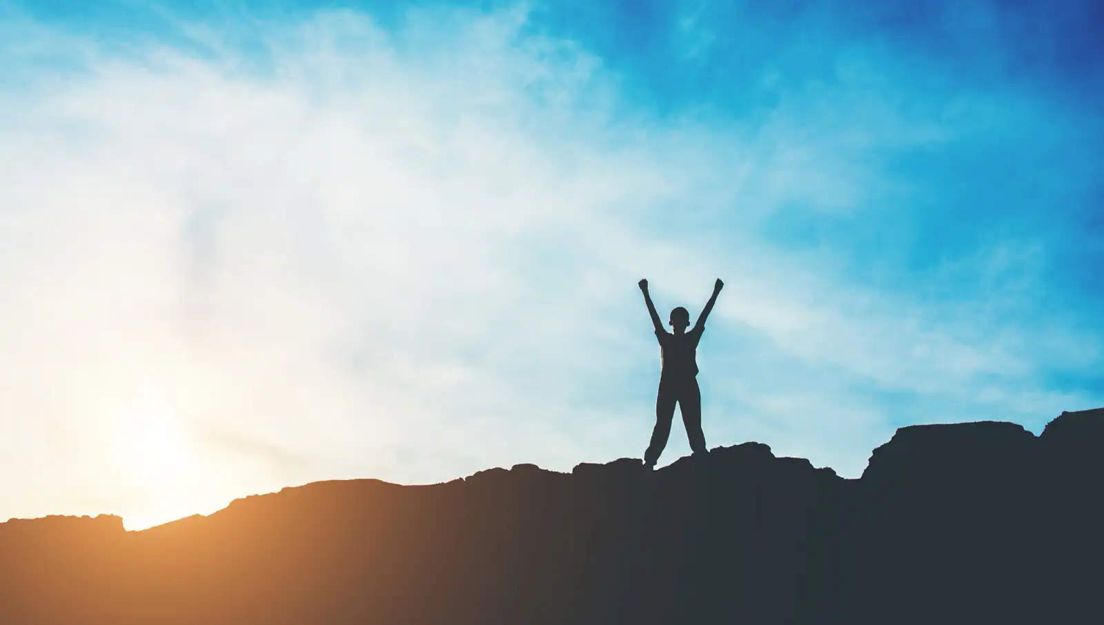 Person standing on a hill at sunrise with arms raised, symbolising success.